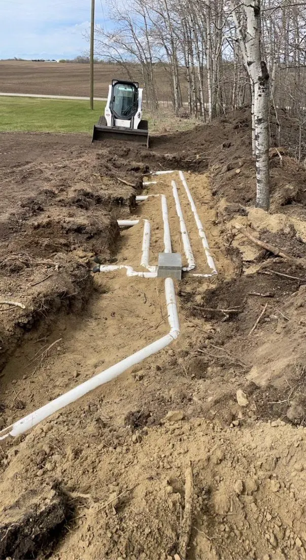 Open trench with new white pipes laid out from a distribution box to the drain field lines, with a skid steer parked at the end of the trench